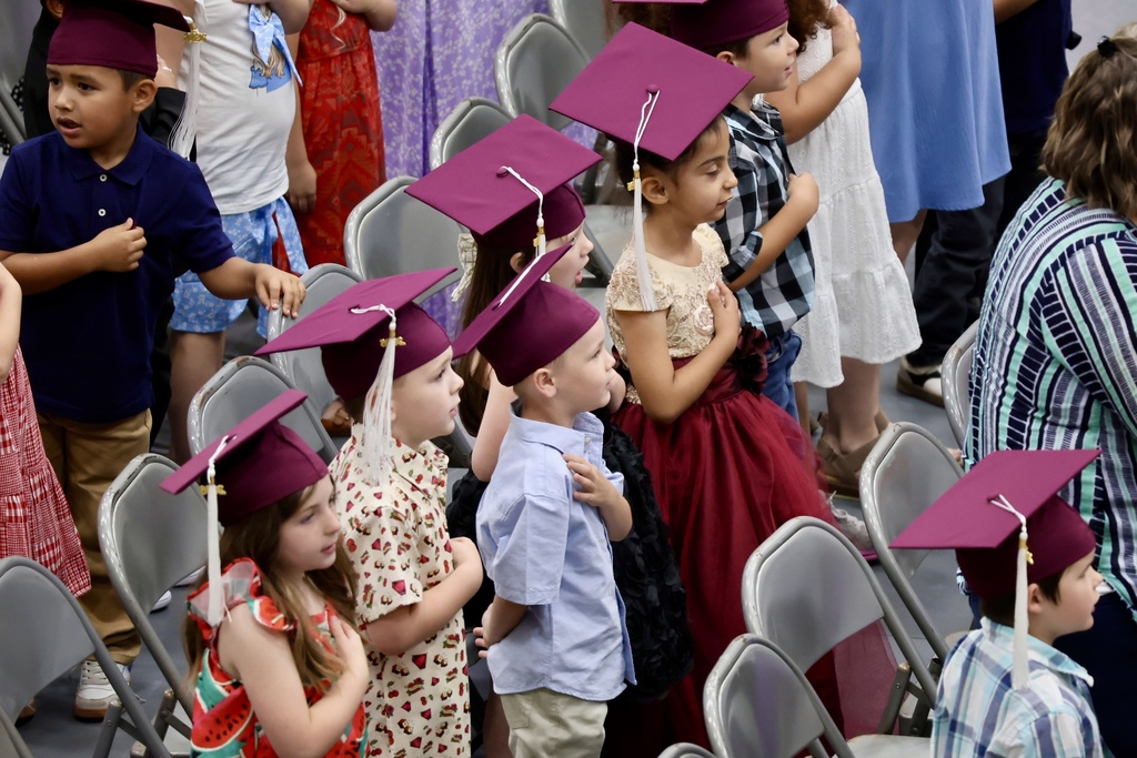 A group of children in caps and gowns standing in chairs, likely at a graduation ceremony.