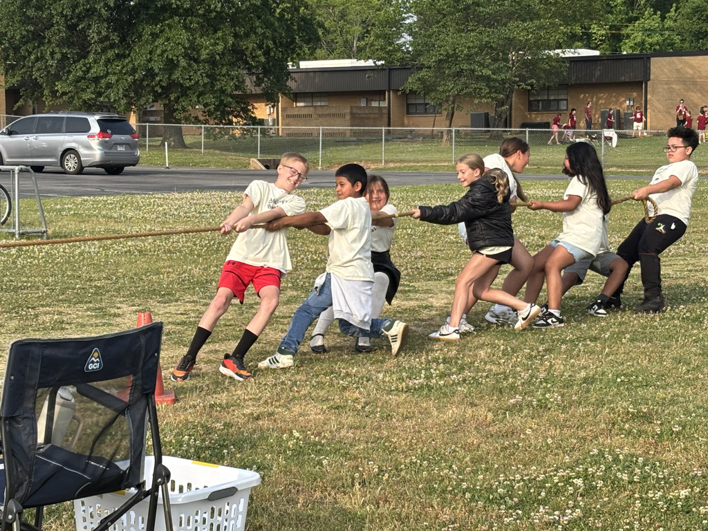 Children participate in a tug-of-war game on a grassy field with a school building in the background.