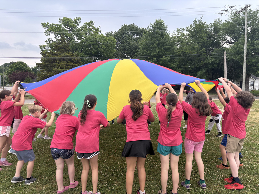 Kids wearing pink shirts stand in a circle holding a colorful parachute. Trees in the background.