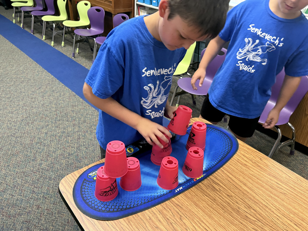 Two children in blue shirts stand at a table, arranging red cups in a pyramid formation on a blue mat.