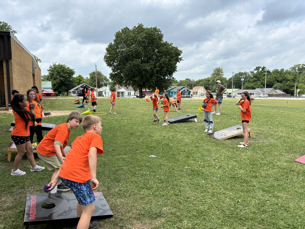 Group playing cornhole outdoors on grass. Some wear orange shirts; a tree and cloudy sky in background.