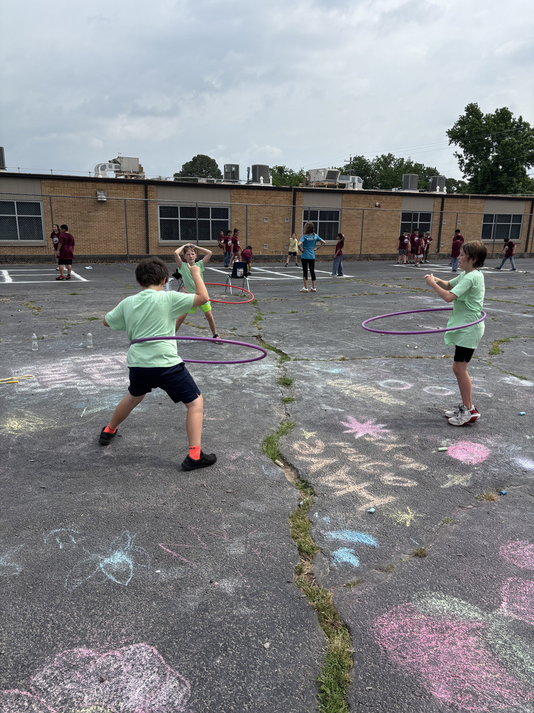 Kids in green shirts hula-hoop outside on a pavement with chalk drawings, a building in the background.