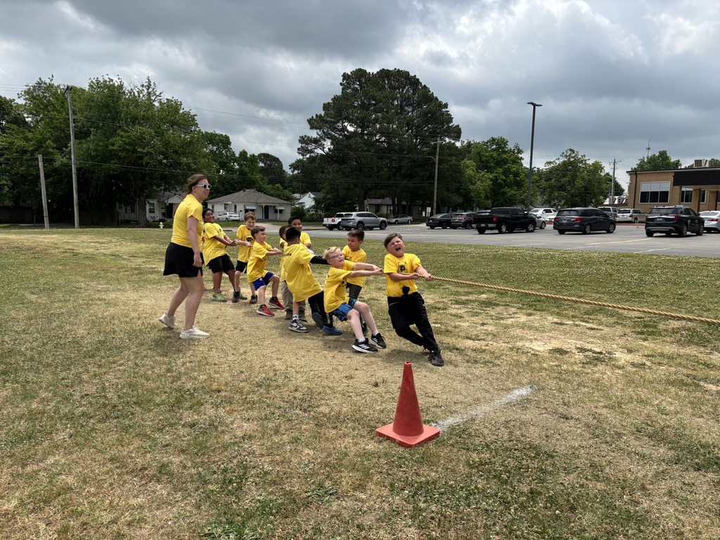 A group of children in yellow shirts play a tug-of-war game on a grassy field, with a traffic cone nearby.