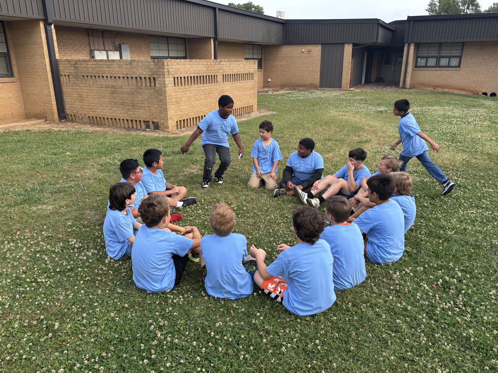 Kids in blue shirts sit in a circle on a lawn, facing an adult standing in the center.