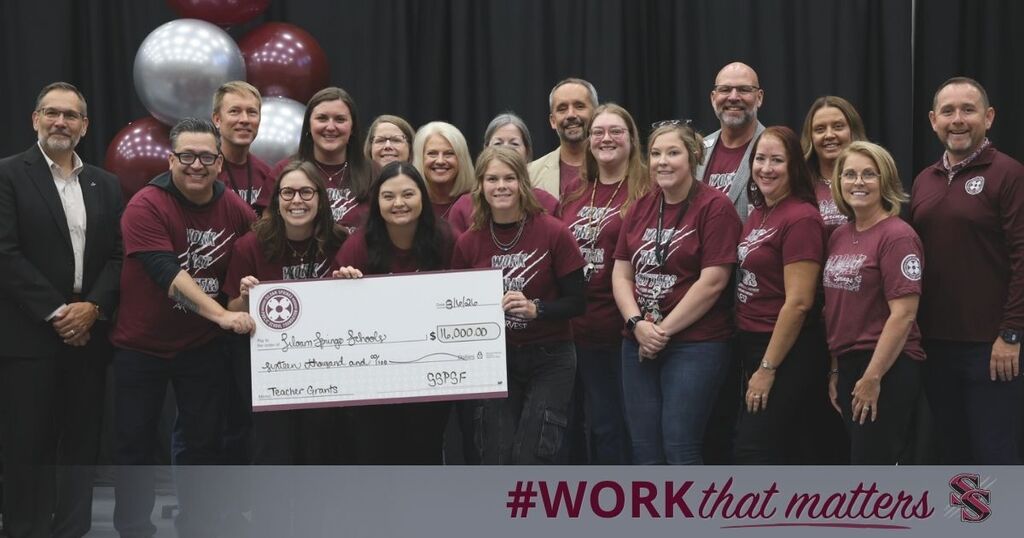 A group of people in maroon shirts holding a large check. Behind them, balloons and a black backdrop.