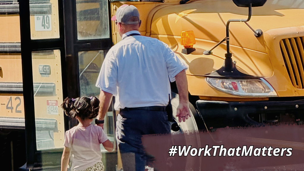 A man in a cap and light blue shirt guides a young girl by her hand near a school bus.