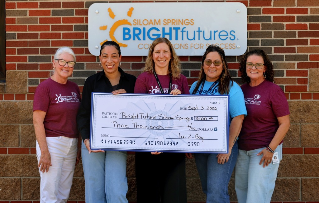 Five women pose in front of a building, holding a large check. They wear maroon shirts.