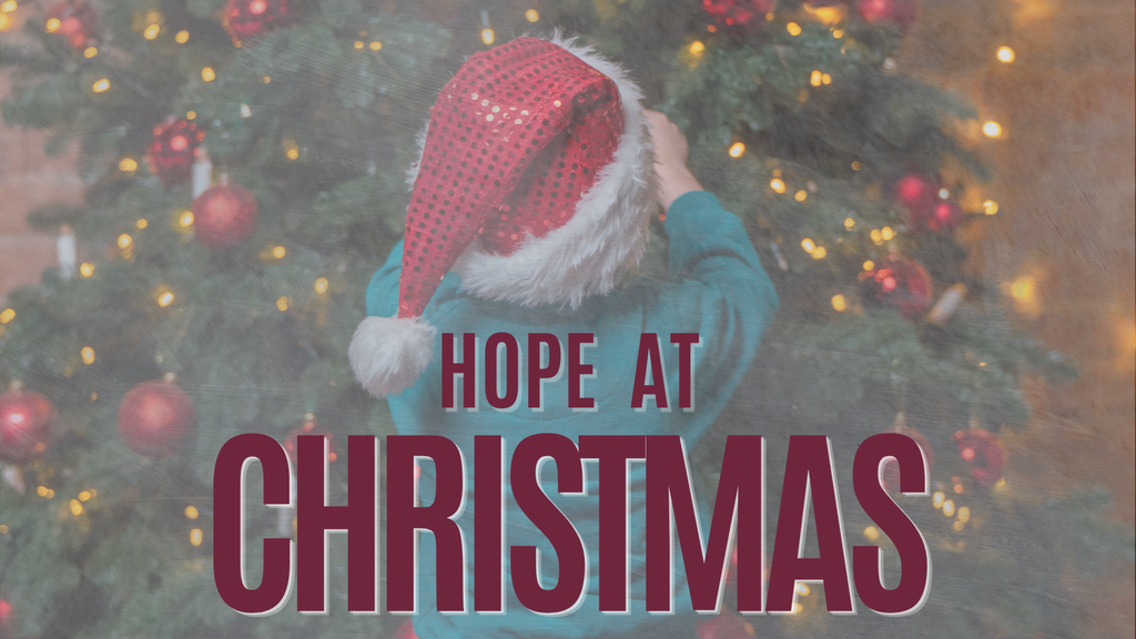 A child wearing a Santa hat decorates a Christmas tree. The tree is adorned with red ornaments and lights.