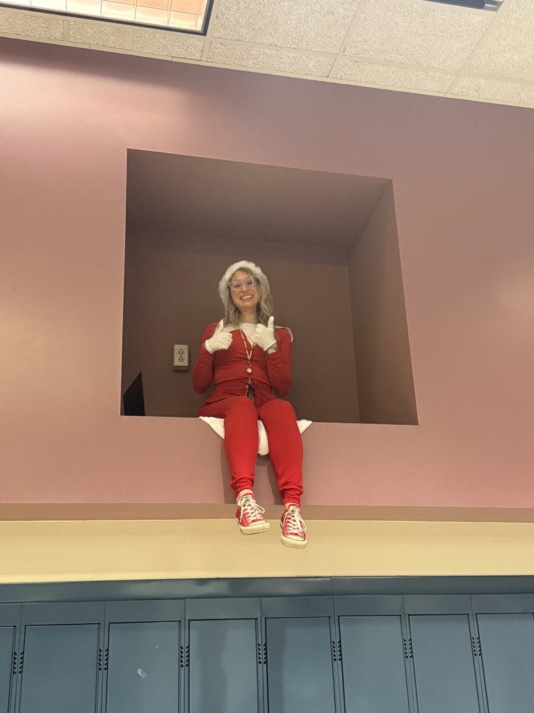 Miss Neubauer dressed as an elf sitting on a high shelf giving thumbs up
