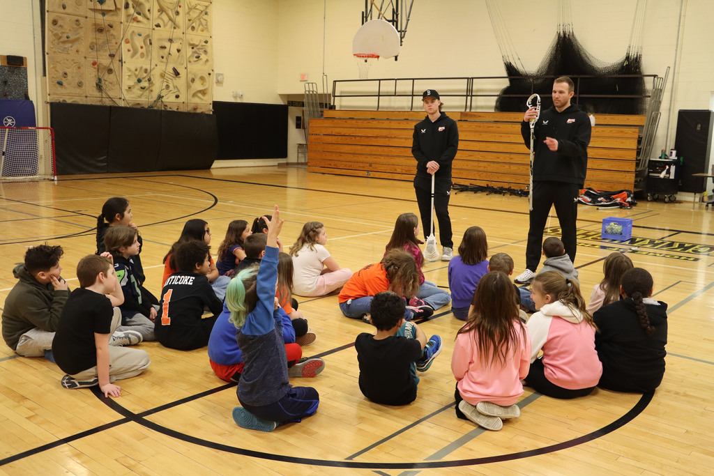 two players holding lacrosse sticks  present to a group of students. 