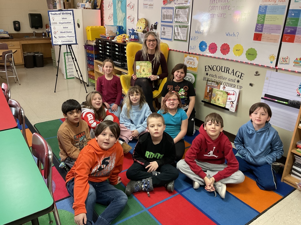 Miss Neubauer holds a book as elementary students pose around her smiling