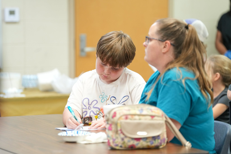 A boy is writing on a piece of paper sitting next to his mom.
