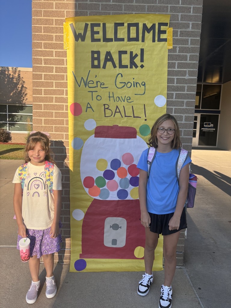 2 students in front of a sign that says "welcome back! we're going to have a ball!" with a picture of a gumball machine