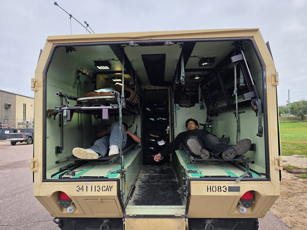 Harry Hopkins Career Academy students explore the interior of a military transport vehicle during a National Guard visit. Several students are seated or lying on the vehicle’s interior stretchers, with the vehicle’s equipment and safety straps visible.