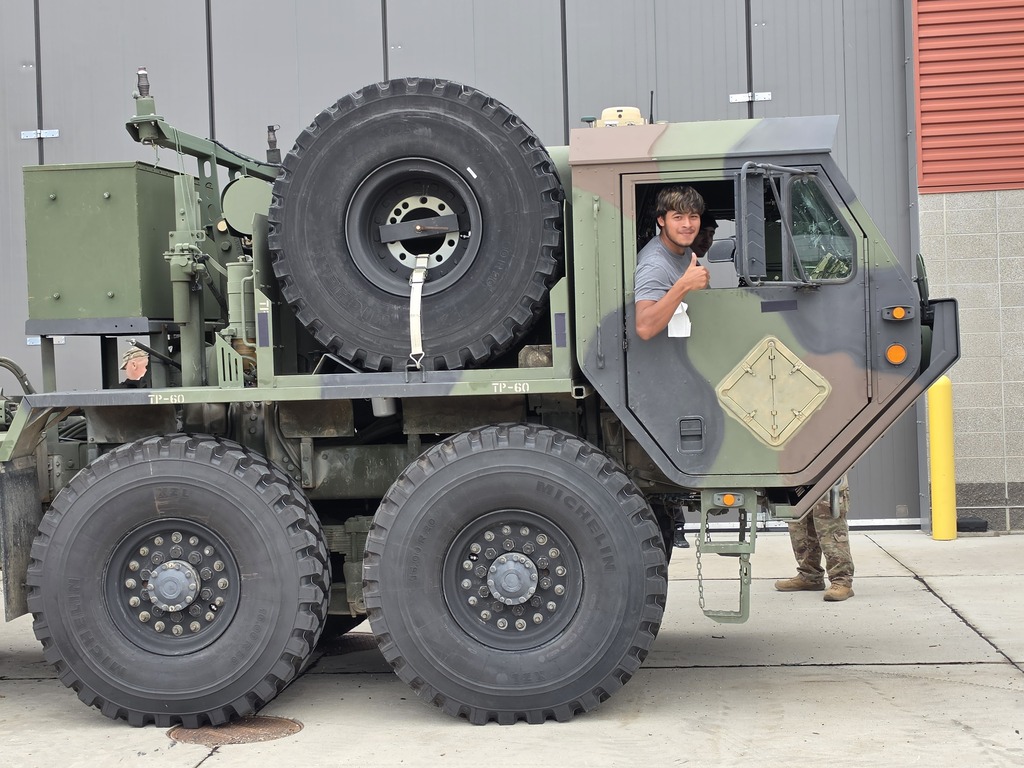 Harry Hopkins Career Academy student sits in the cab of a large military vehicle and gives a thumbs-up while participating in a National Guard visit. Another service member is visible near the vehicle, which is parked outside a school facility.