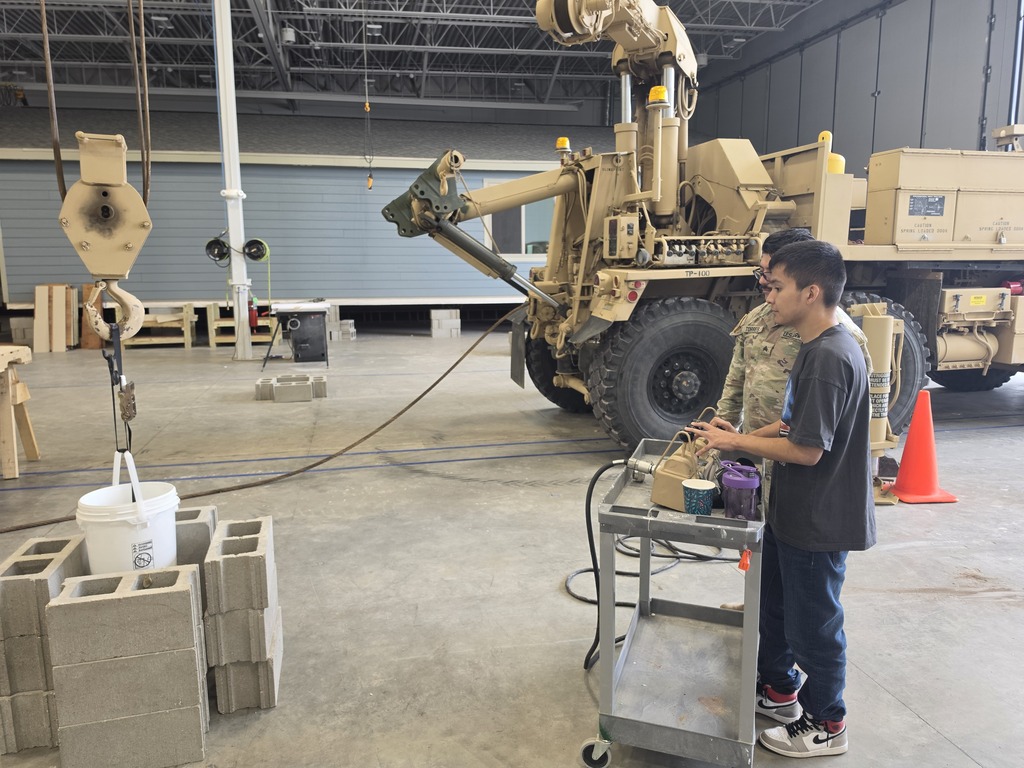 Harry Hopkins Career Academy student operates a control panel beside a large military vehicle inside a National Guard facility, while a service member observes. A heavy-duty lifting mechanism, equipment, cables, and safety cones are visible throughout the workspace.