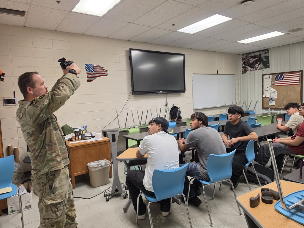 National Guard service member demonstrates equipment to a group of Harry Hopkins Career Academy students in a classroom. Several students sit at tables and watch the demonstration, while classroom tables, chairs, a display screen, and American flag decorations are visible.