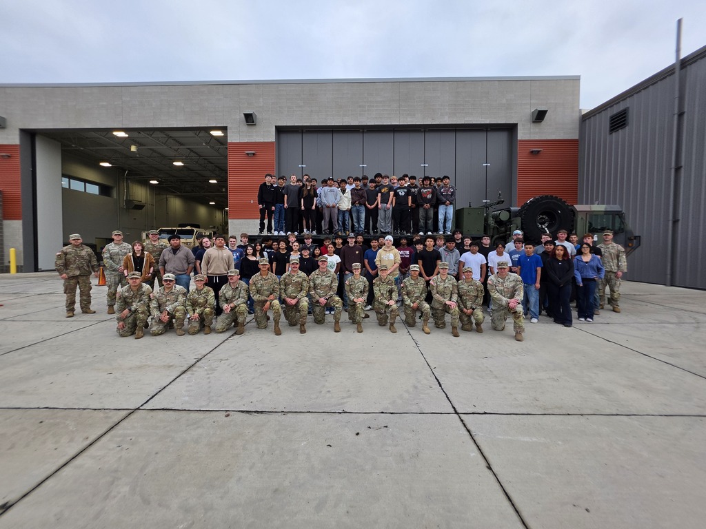 Large group photo of Harry Hopkins Career Academy students and National Guard members gathered outside a school facility with military vehicles in the background. Students stand in several rows behind National Guard members in uniform, who are seated and standing in front.