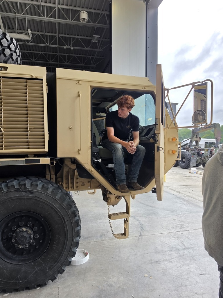 Harry Hopkins Career Academy student sits in the driver’s seat of a large military vehicle during a National Guard visit. The vehicle’s large tire, cab, steps, and surrounding equipment are visible inside the facility.