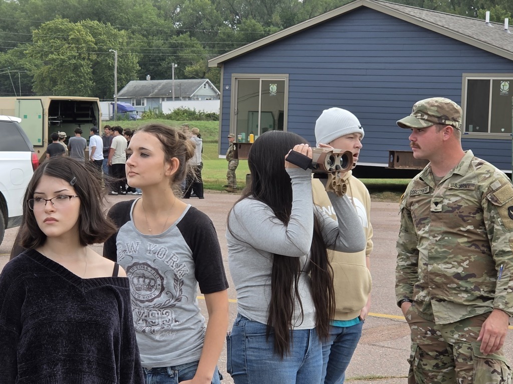 Harry Hopkins Career Academy students participate in a National Guard demonstration outdoors. A student looks through a handheld optical device while a National Guard service member in uniform observes nearby. Other students and a military vehicle are visible in the background.