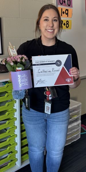woman with dark stands in front of white board holding flowers and certificate