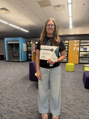 woman with light hair stands in classroom holding a green and white certificate