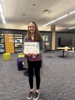 woman with light hair stands in classroom holding a green and white certificate