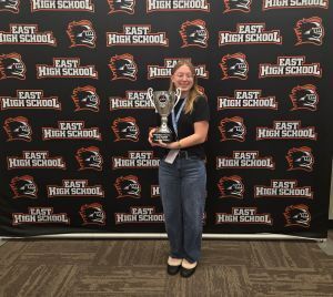 woman holds a trophy while standing in front of a black and orange East High School  backdrop