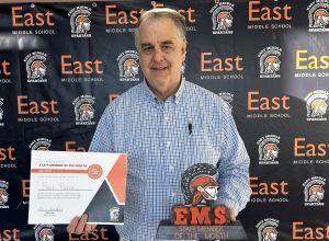man with gray hair stands in front of black and orange East Middle School backdrop