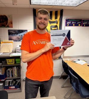 man in orange shirt stands in classroom holding up a certificate