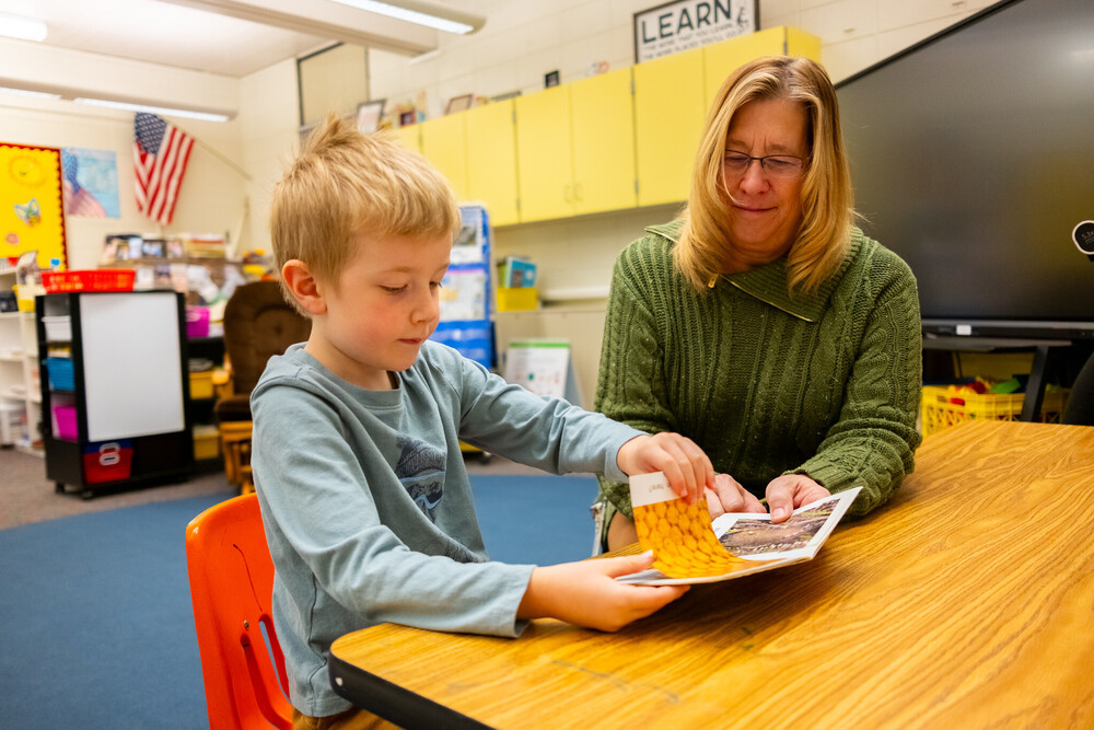 Woman smiles happily as she sits next to a young reader looking at a book