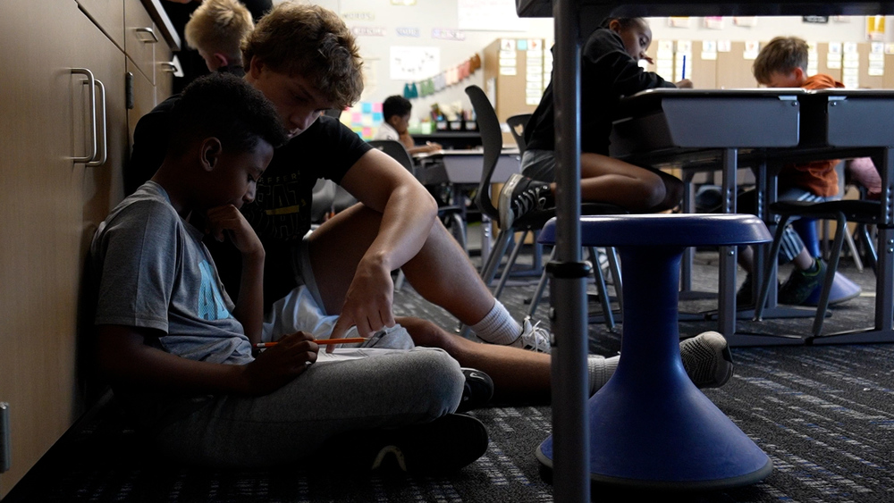An older student sits next to younger student against a cupboard. The older student helps the younger one with school work and points at the paper while other young students work at desks in the background