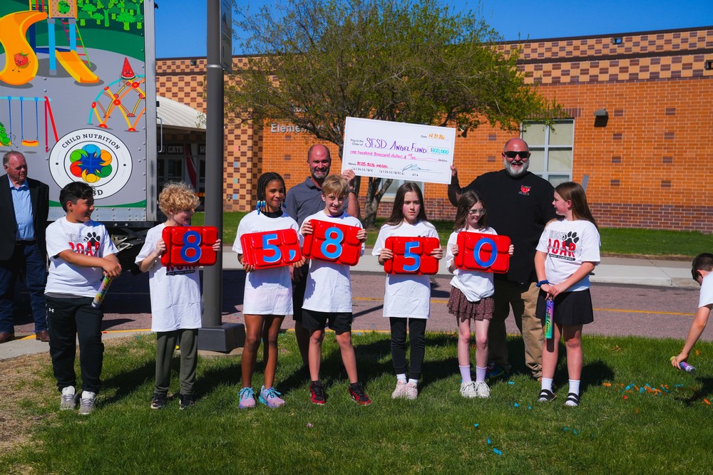 A group of children hold up school lunch trays with the number 85,850 on them. Behind them, 2 men hold a big check writen to the the SFSD Angel fund in the amount of $100,000. 