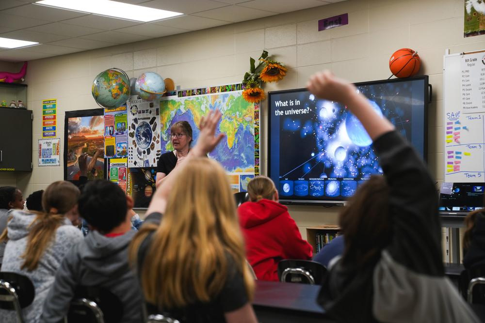 Students raise their hands during a presentation with an educator at the front of the classroom. A screen says "What is in the Universt?" in the background.