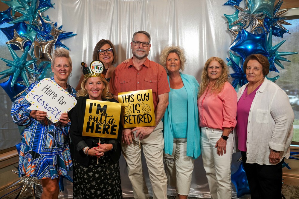 Group of educators, including Ben Reifel Middle School retirees, Barb Christophersen and Todd Peichel stand in front of a photo backdrop smiling with one another holding funny signs like “I’m outta here” and “This guy is retired” and “Here for the dessert” at the Annual 2026 Recognition Banquet. 