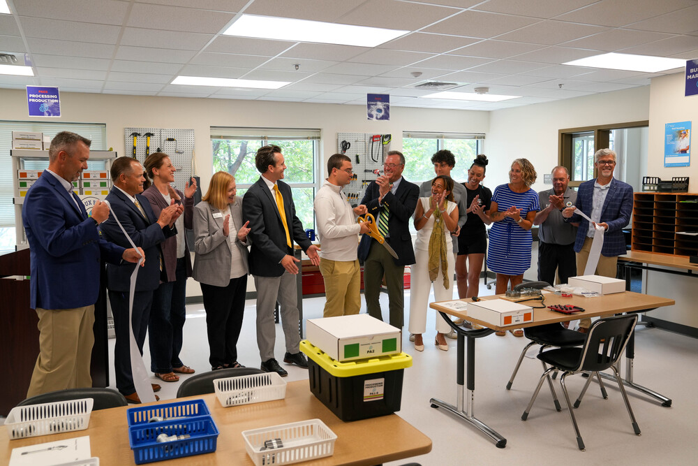 Ribbon cutting in a classroom. 13 people are clapping and smiling while a young man in the middle holds a large pair of scissors, having just cut the ceremonial ribbon.