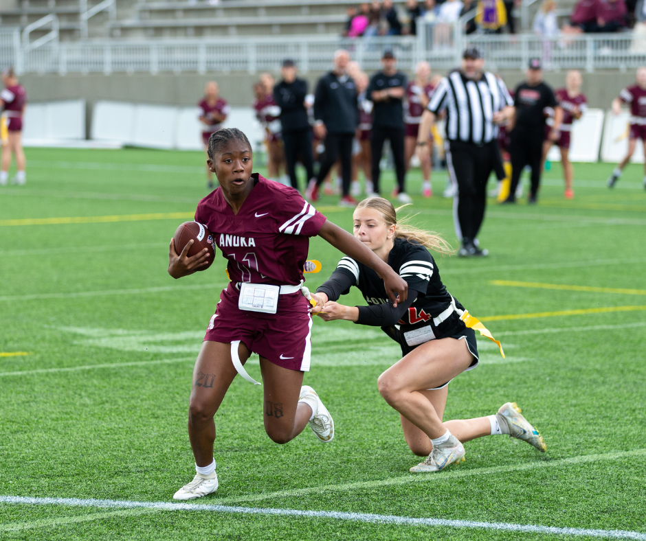 Young woman strongly defends and holds on to a football on the field while an opposing player tries hard to grab a flag from her belt