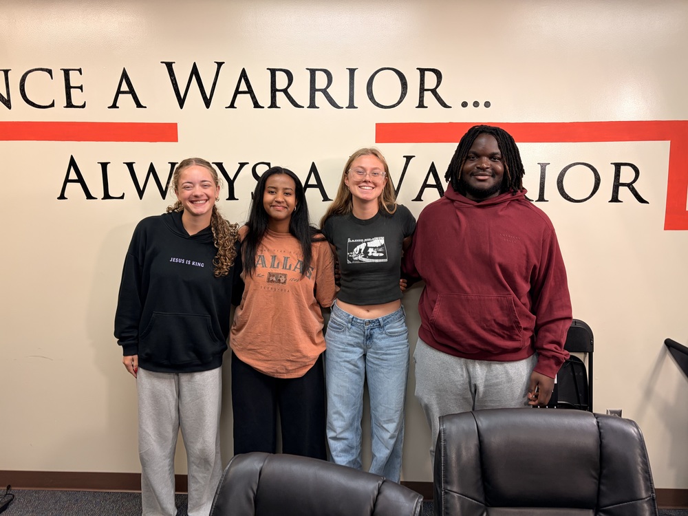 Four young students stand in front of a "once a warrior, always a warrior" mural smiling at the camera