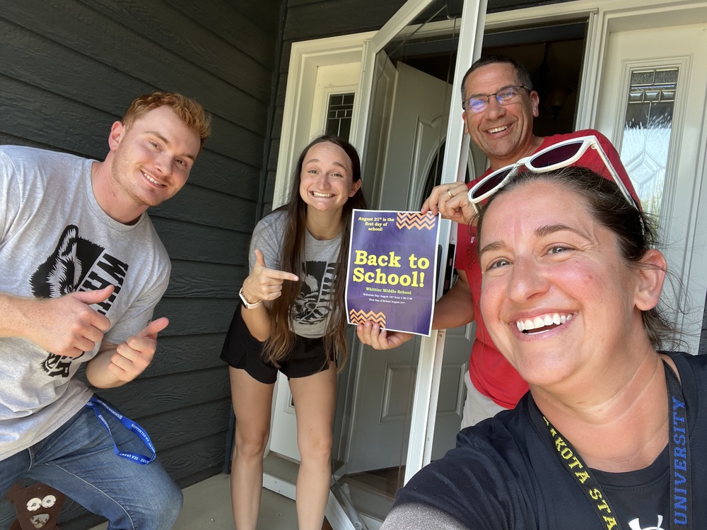 four individuals stand in front of an open front door with excited smiles on their faces holding a back to school sign