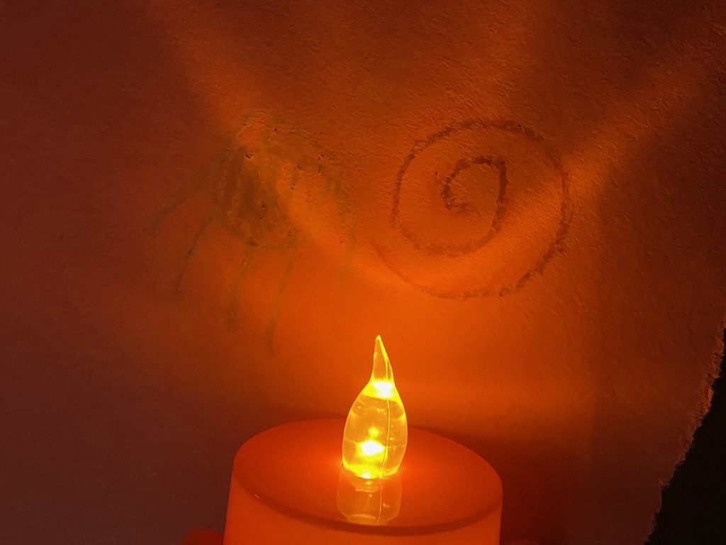  A close-up of a flameless, orange LED candle casting a warm glow on a piece of textured, brownish paper. A simple swirl and other faint marks have been drawn on the paper in the limited light.