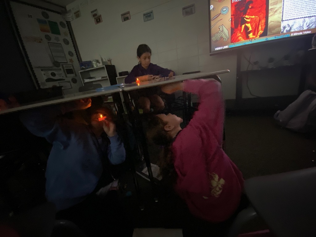 A wide view of a darkened classroom where students are recreating cave art. Two students are on the floor, looking up and drawing on paper taped underneath a desk, using flameless candles for light. Another student is visible in the background.