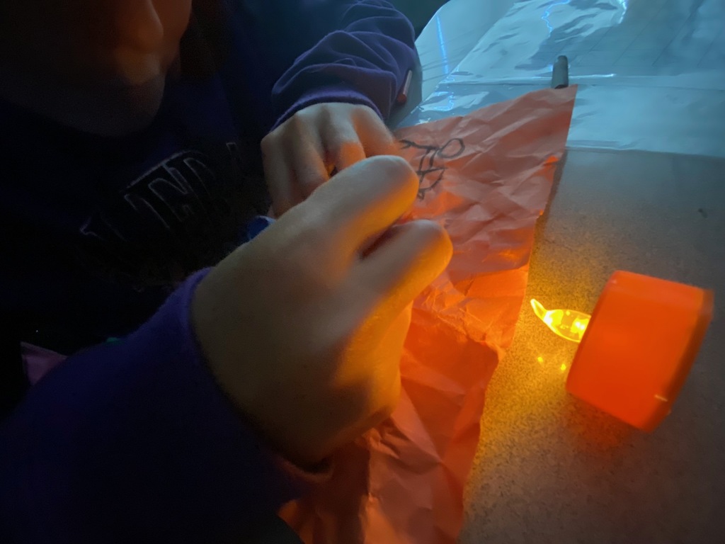 A close-up of a student's hands drawing on a piece of crumpled orange paper. An orange, flameless candle sits beside their hand, illuminating their work and showing the beginnings of a simple line drawing.