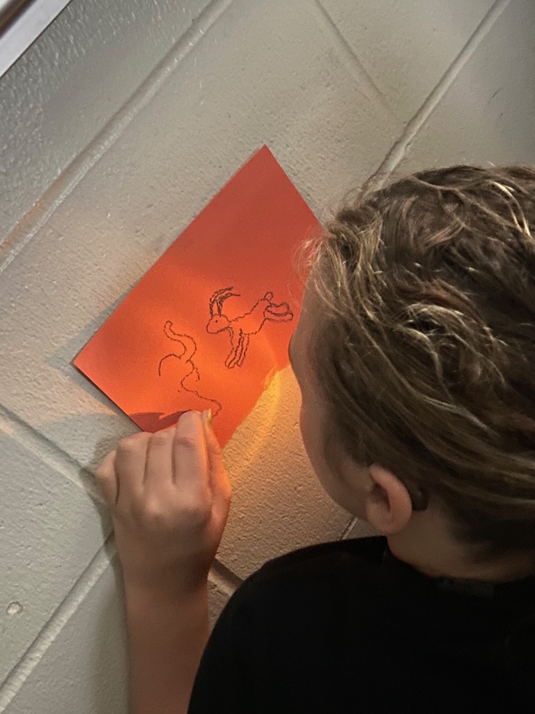 An over-the-shoulder view of a student examining their cave art drawing on a sheet of orange paper. The drawing, illuminated by a flameless candle, depicts a goat-like animal and a winding line.