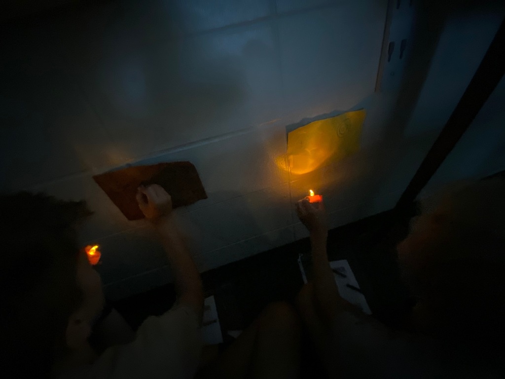 An atmospheric, dimly lit shot showing two students from behind as they draw on paper taped to the underside of a surface, mimicking a cave ceiling. Each student holds a flameless candle up to their paper to see their work.