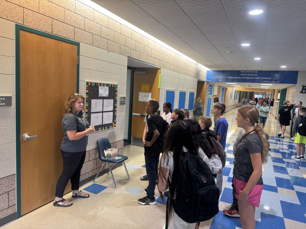 A female teacher stands in a busy school hallway next to a game board mounted outside a classroom door. A large, engaged group of middle school students is gathered around her, looking at the board as they participate in the math game.