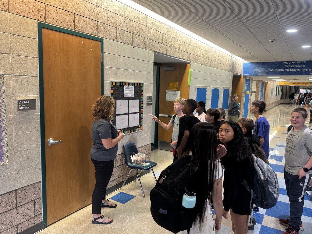 A female teacher stands in a busy school hallway next to a game board mounted outside a classroom door. A large, engaged group of middle school students is gathered around her, looking at the board as they participate in the math game.