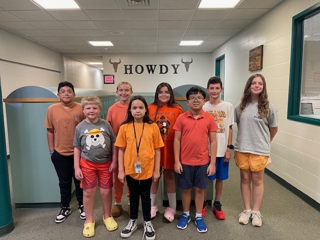 A large group of smiling middle school students stand together in a school hallway, all wearing orange for Orange Shirt Day.