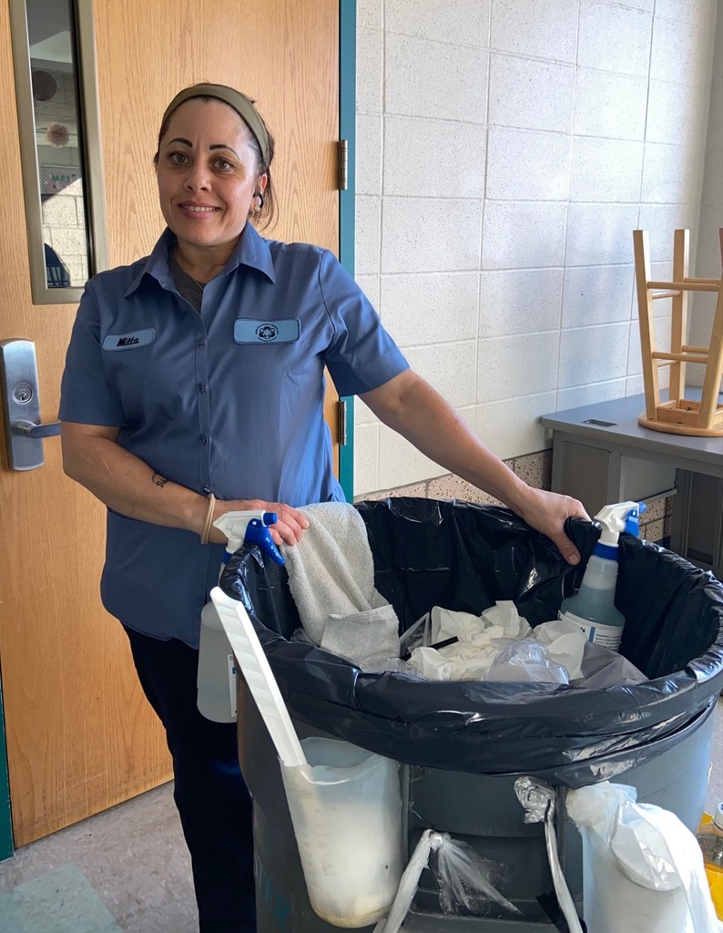 Custodial staff member stands at a classroom doorway beside a gray rolling trash can and cleaning bottles, smiling; stacked stools are visible behind.