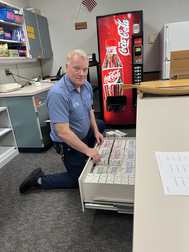 Maintenance staff member kneels to organize a drawer filled with tissue boxes in a staff workroom; a Coca-Cola vending machine and small U.S. flag are in the background.