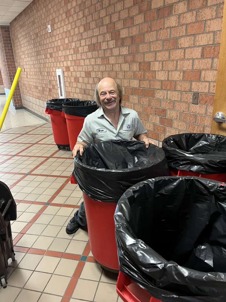 Custodial staff member smiles while replacing a trash liner in a large red rolling bin in a school hallway with brick walls and tiled floor.